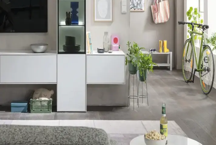 Wohnbereich mit weissem Lowboard und hohem Vitrinenschrank, davor Couchtisch mit Flasche und Schale, rechts Flur mit grünem Fahrrad, Bank und vielen Pflanzen.